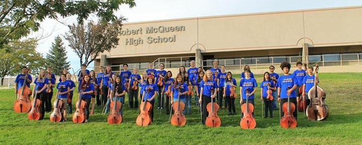 High school students standing in three rows wearing matching t-shirts and jeans on the lawn in front of their high school. Each student is holding their instrument, a violin, viola, cello or upright bass in front of them.
