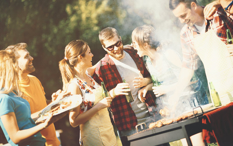 Group of men and women laughing and holding plates of food next to a grill in a backyard.