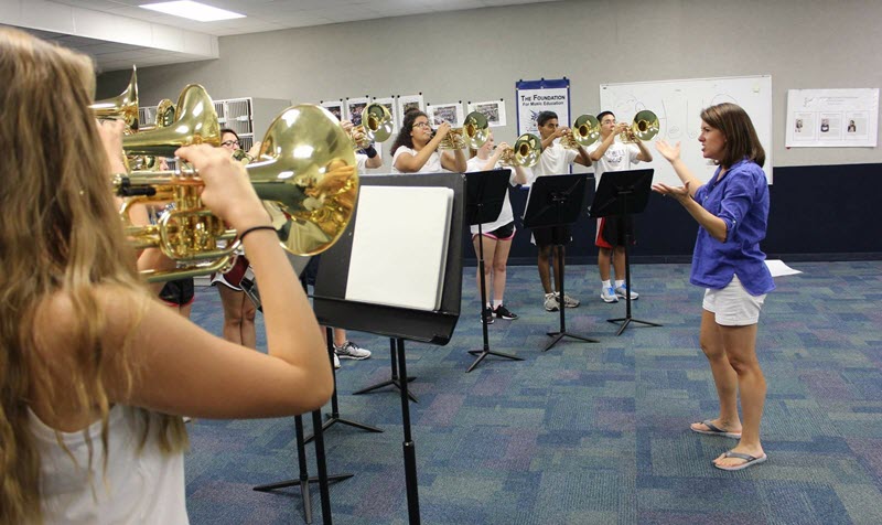 Female band teacher conducts practice with brass players in a large room with carpet.