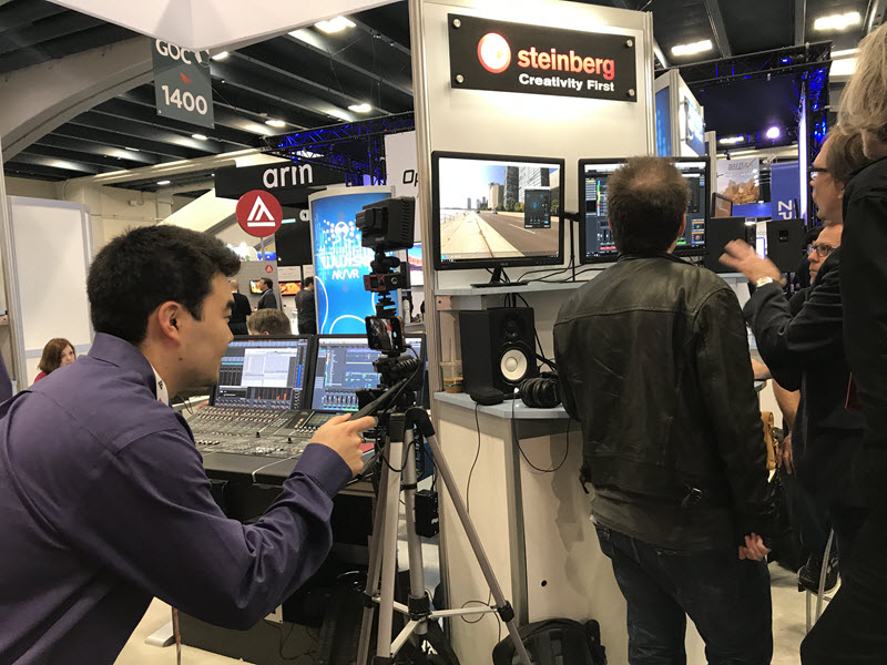 A man video recording two other men at a tradeshow booth. One of the subjects in booth is using his hands and speaking to indicate something that is happening on computer screens hanging on booth wall.