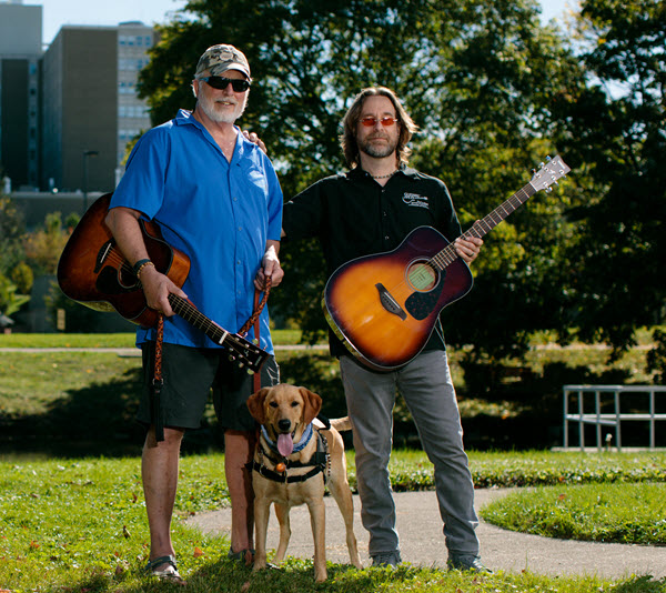 A man with a service dog standing next to another man in a park. Both men are holding acoustic guitars.