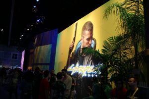 Large crowd clustered in front a large wall where a graphic image of a young man holding a gun and the title "CyberPunk 2077" is emblazoned.