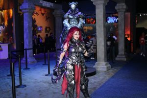 Young woman dressed in a costume emulating the futuristic armor and weaponry of the "Darksiders Genesis" statue behind her.