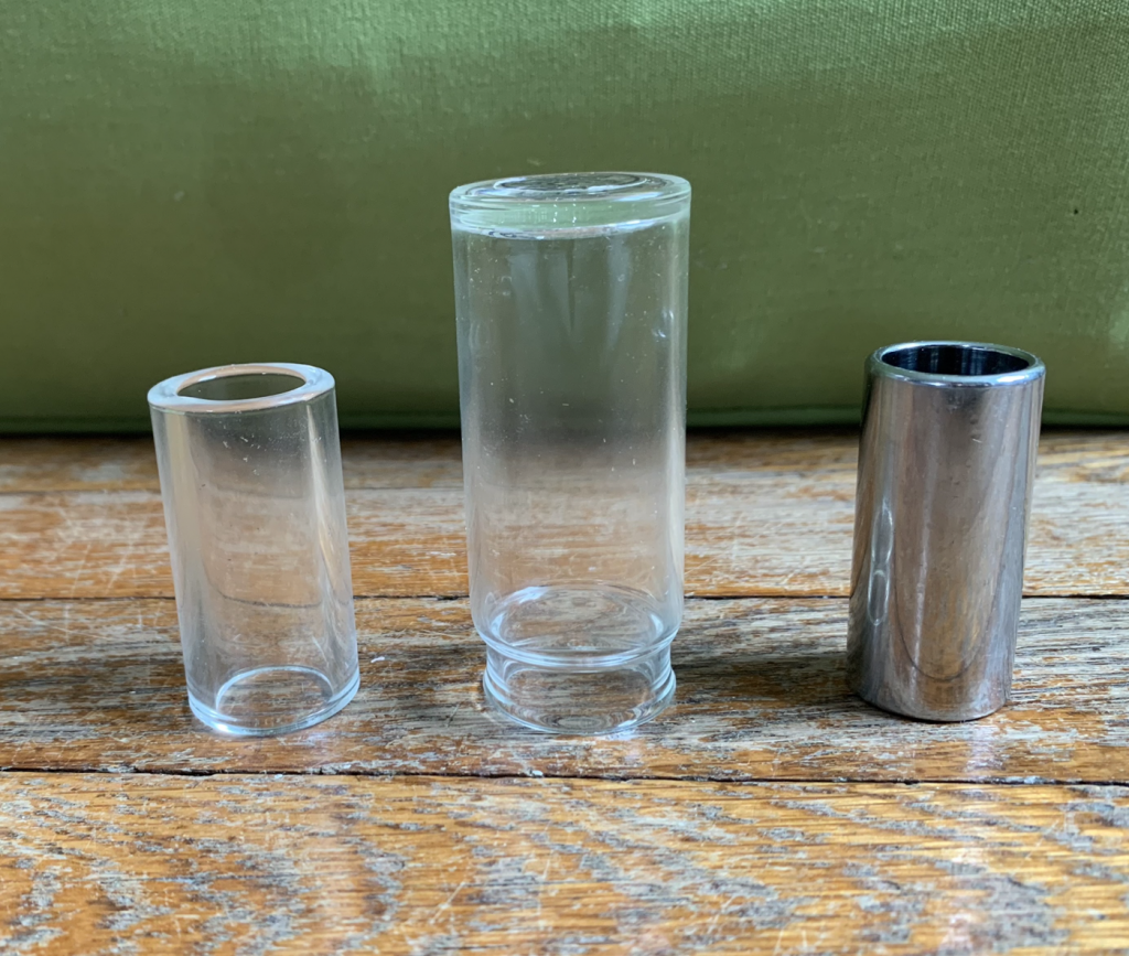 Three slides for playing slide guitar are lined up on a table. From left to right, there is a small open-ended glass tube, a larger closed-ended glass tube, and a small open-ended metal tube.