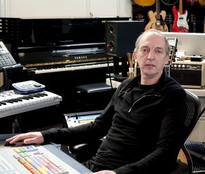 Middle aged man behind a sound board and surrounded with studio equipment.