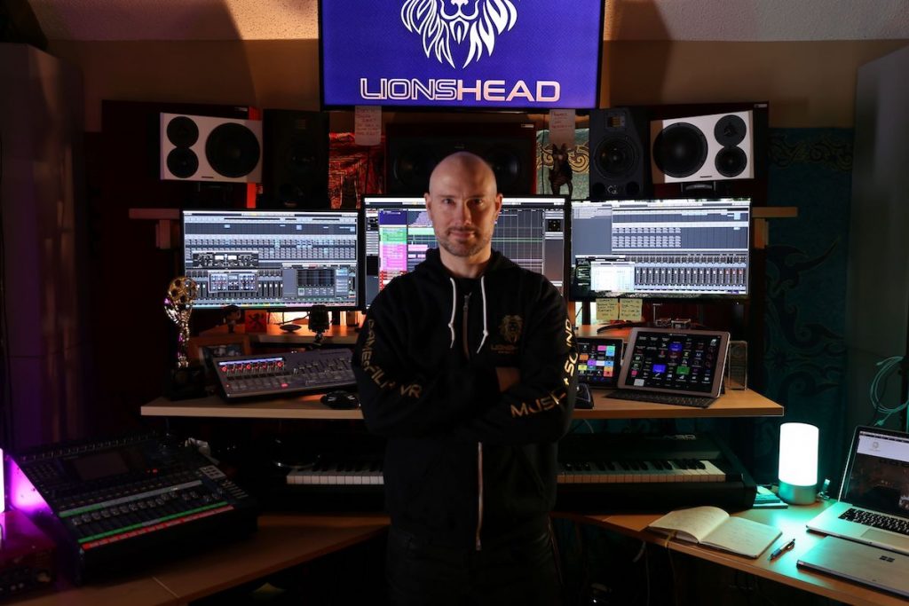 Man in black sweatshirt stands in front of large monitors in sound design studio.