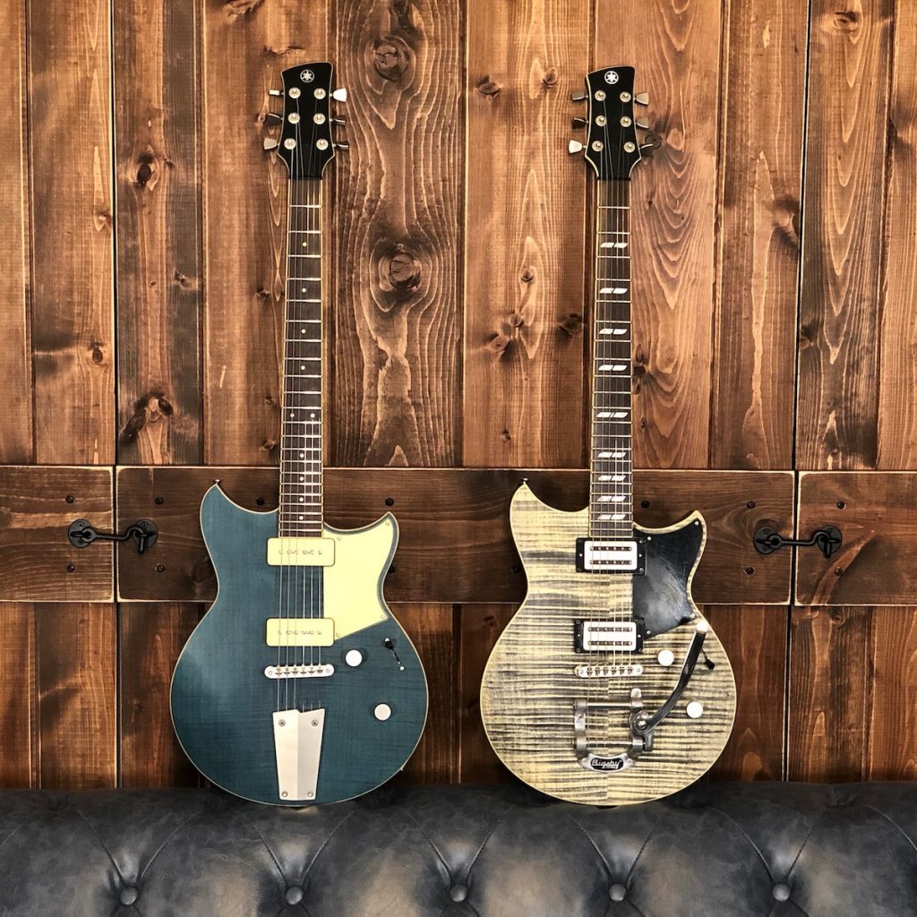 Two electric guitars resting on leather couch with wooden backdrop.