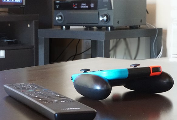 View of a gaming controller and remote control on a table top with an AV receiver in the background.