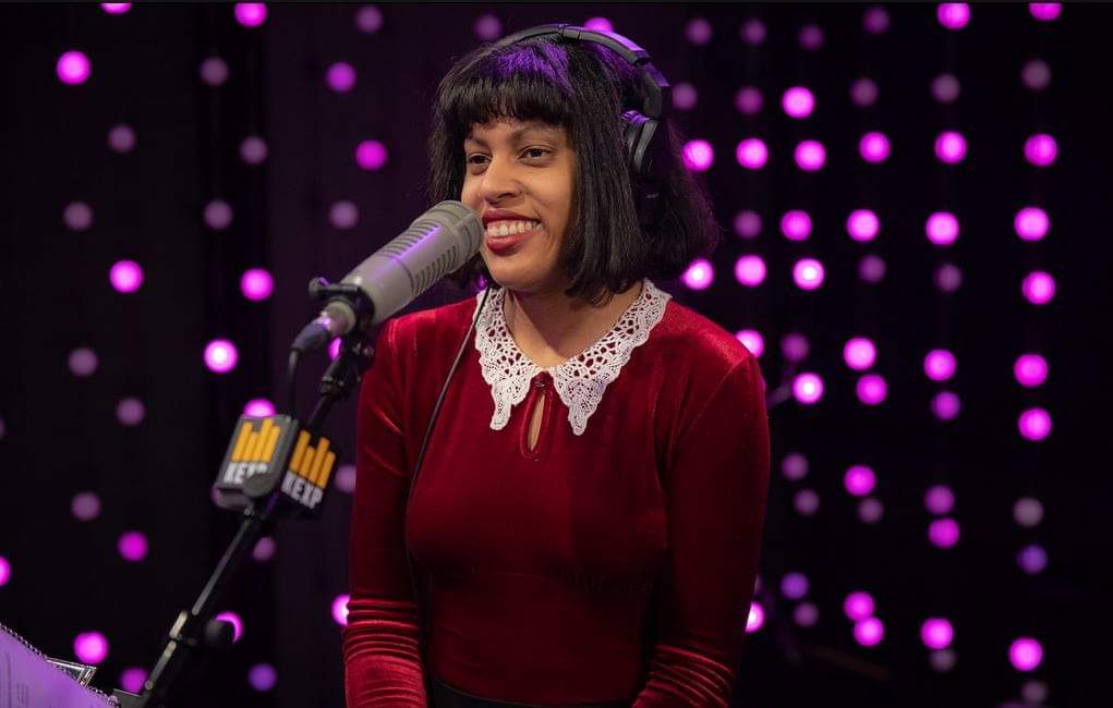Young woman wearing headphones and speaking into a microphone with the call sign of KEXP on it.