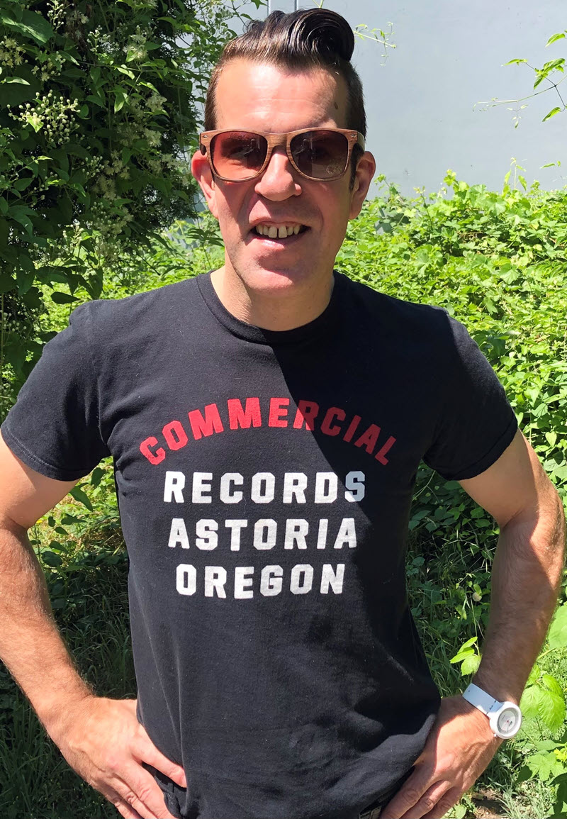 Man in an outdoor setting. He's wearing sunglasses and a t-shirt with the words "Commercial Records Astoria Oregon" on the front of shirt.