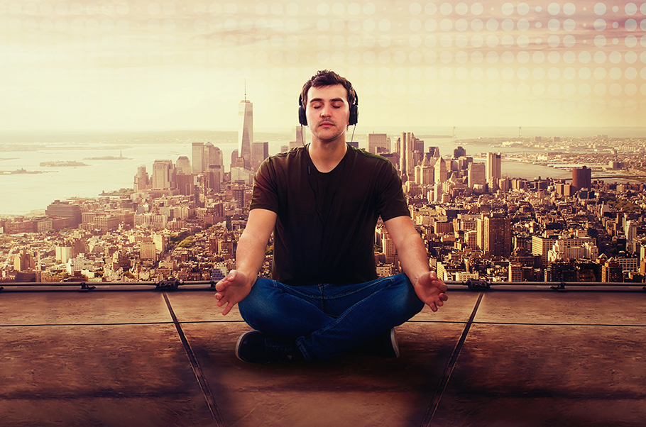 Man sitting in a lotus position meditating on on a roof with a cityscape behind him. He is wearing headphones.