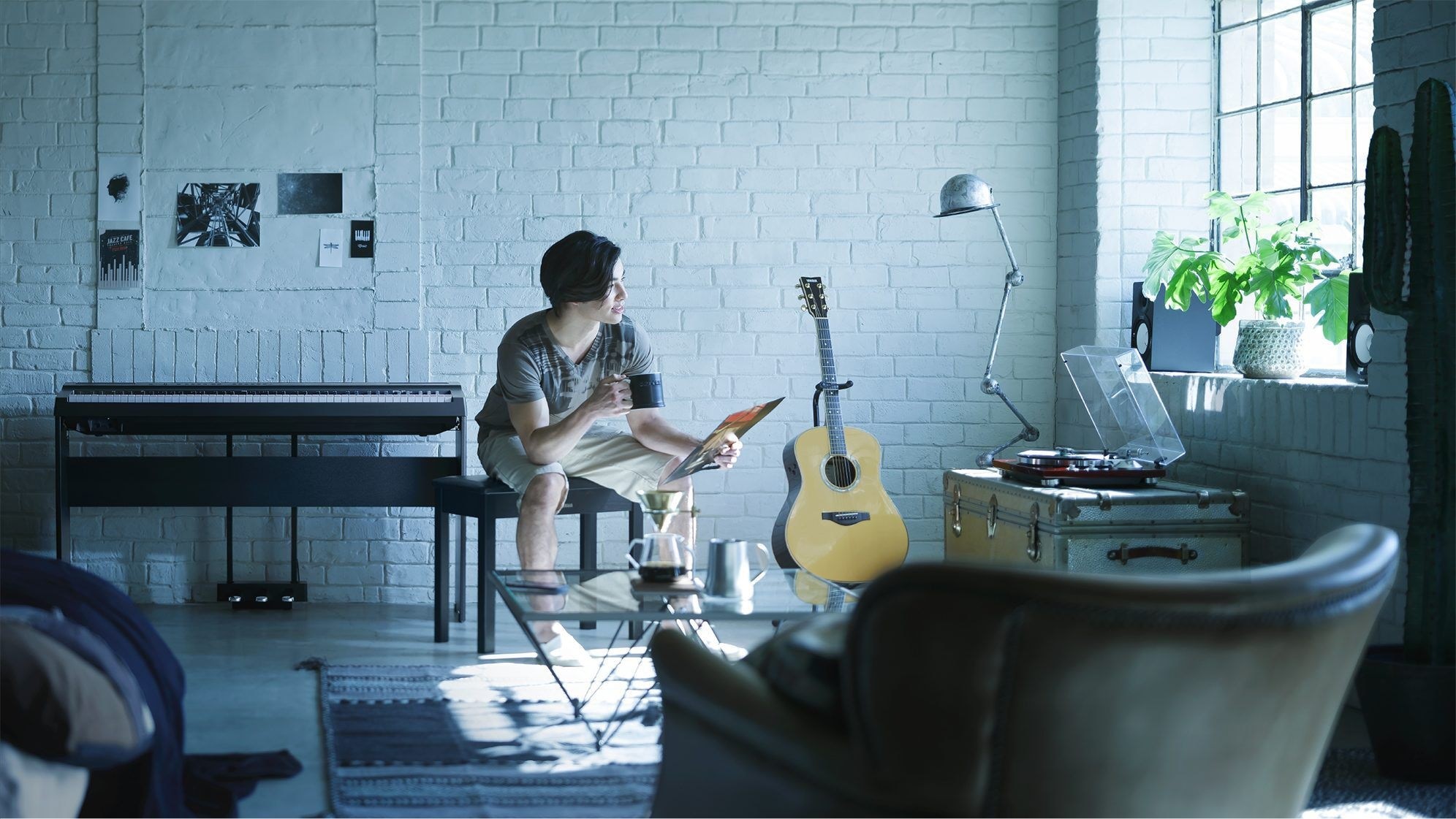 A young adult in a loft-style living room surrounded by an electronic keyboard, a guitar on a stand and amplifier.