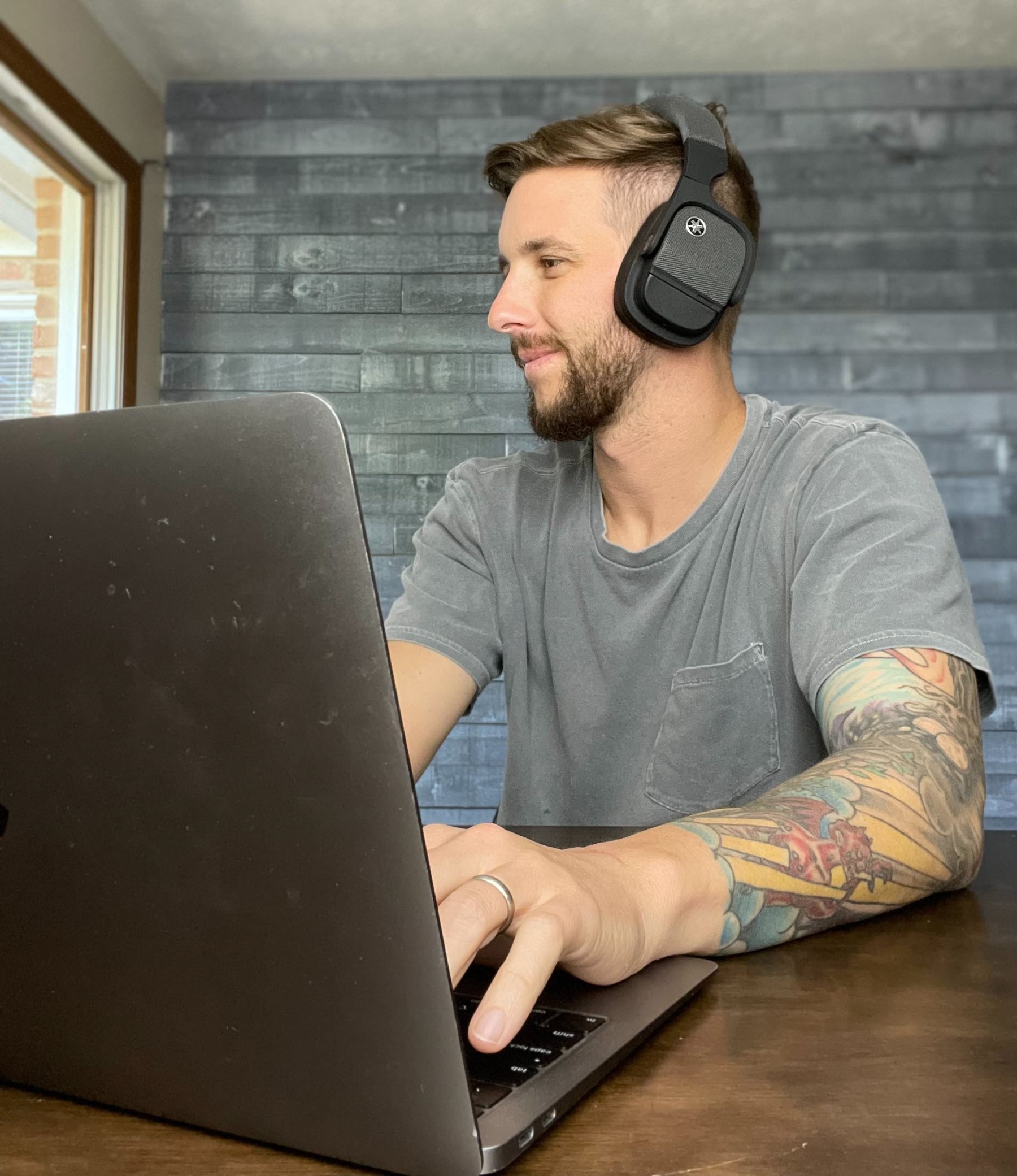 Man in his 30's with facial hair and tattoos on his lower arms is in front of a laptop wearing Yamaha wireless headphones.