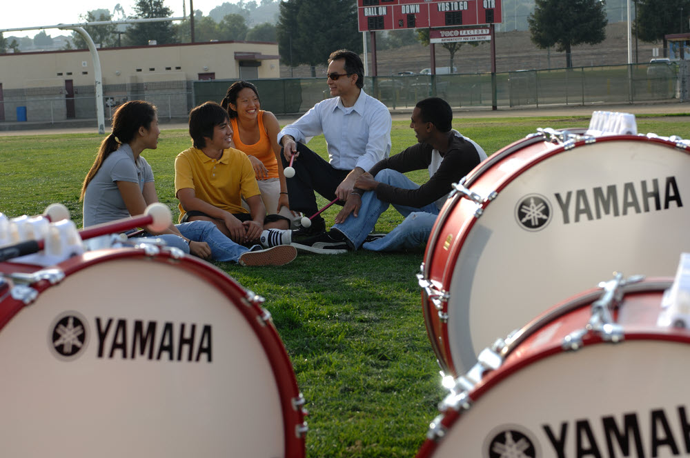 Group of drumline students sitting on grass in background with teacher and bass drums with Yamaha logo n foreground.