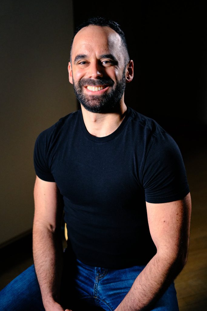 Man in black t-shirt and jeans smiling for camera.