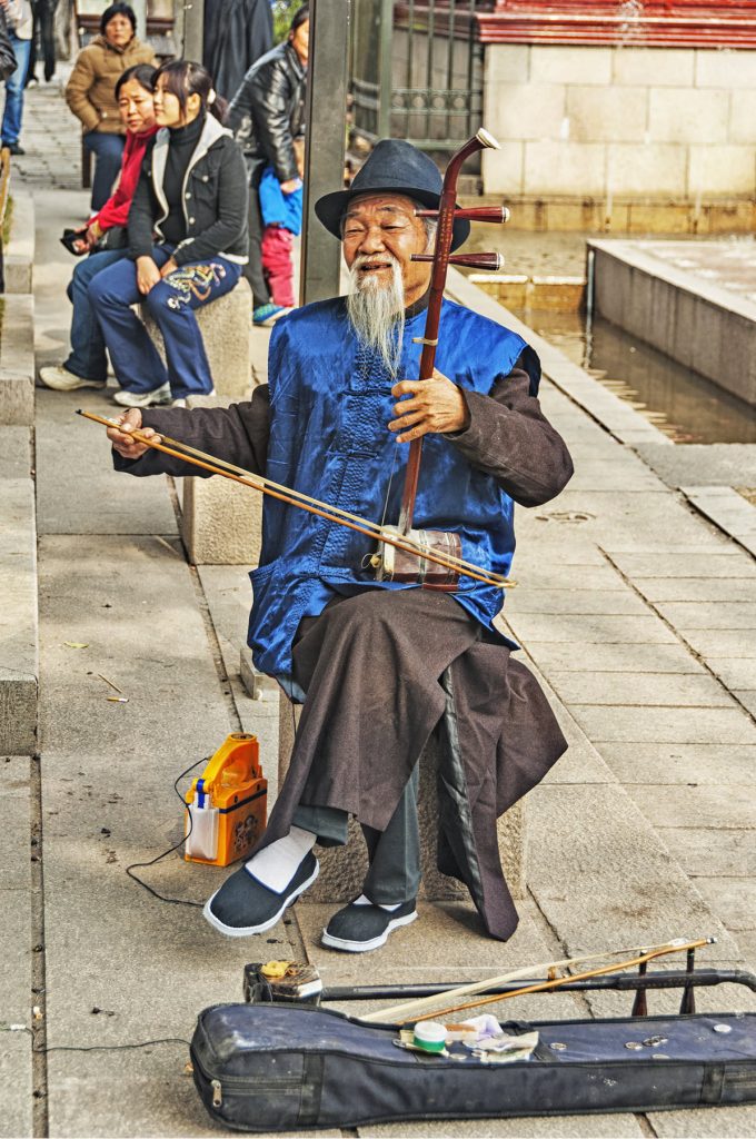 Elderly man with long beard in traditional dress sitting in a plaza playing a stringed instrument with a bow.