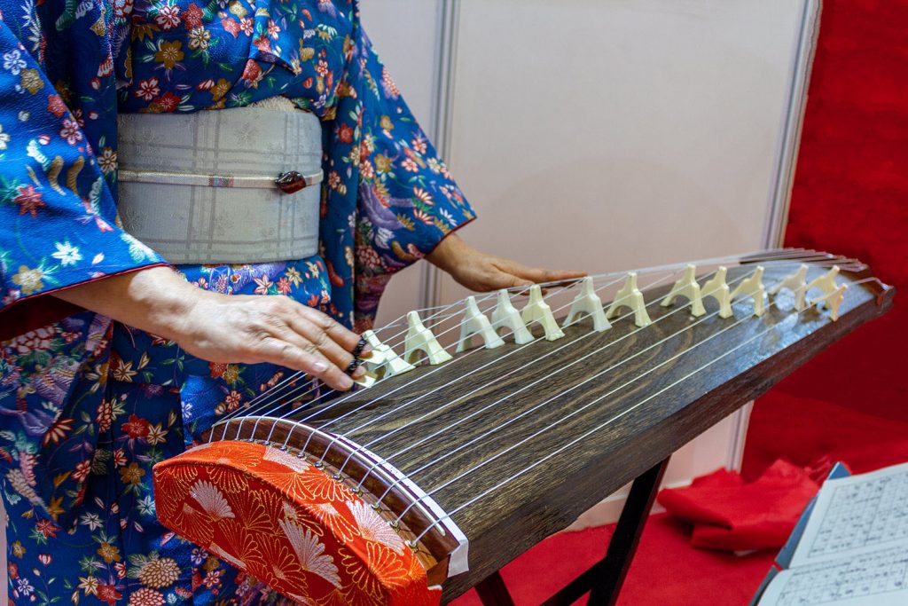 Closeup of a woman in kimono with a long rectangular stringed instrument across her lap. She is playing the strings with her right hand.