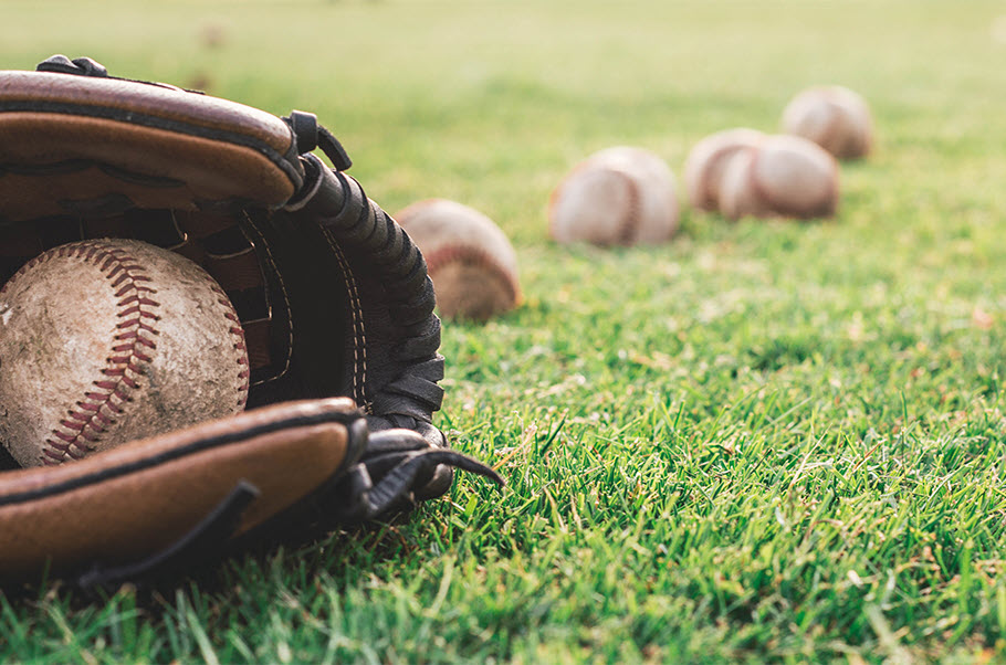 In foreground on the grass are a well-used baseball glove with a dirty baseball inside. In background, there are several baseballs laid haphazardly on the grass.