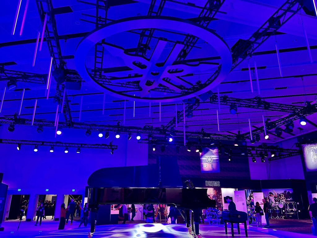 Beautiful grand piano under a theatrical lighting scheme at a tradeshow with drop lights hung from an oversized representation of the Yamaha logo from above. Tradeshow visitors can be seen in background.