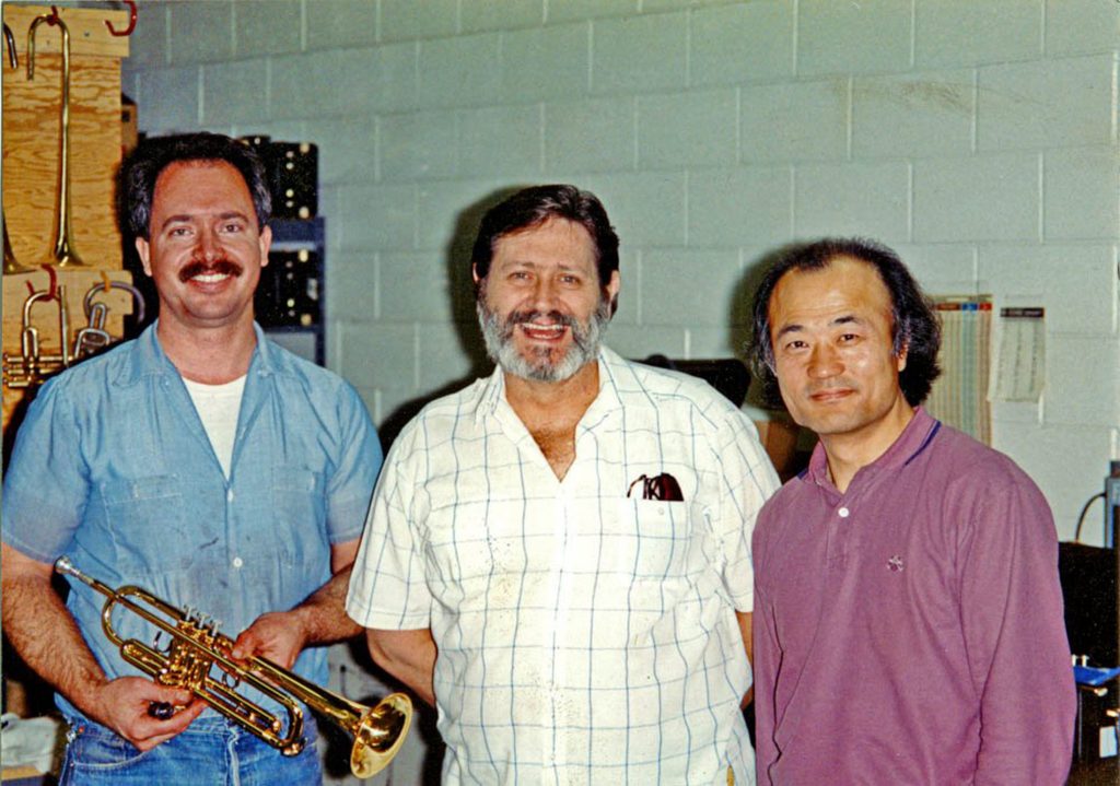 Three men standing side-by-side in a workshop smiling for camera.