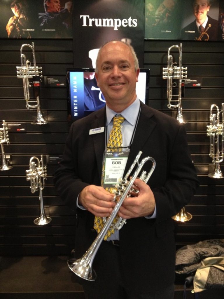 Man in suit smiling for camera while holding a silver colored trumpet. Behind him is an large wall display case of trumpets.