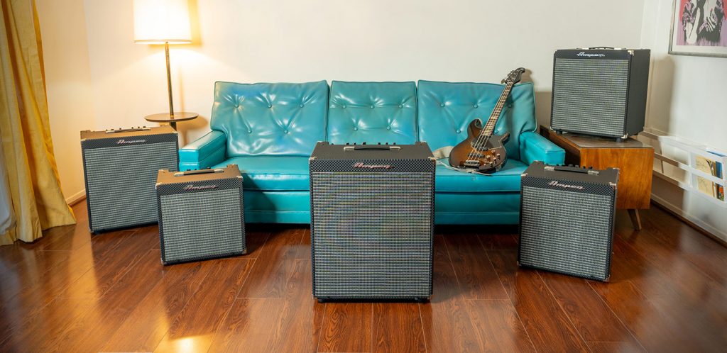 A living room setting with simple floor lamp, hardwood floors and large turquoise couch with bass guitar on it in background. In foreground are a variety of sized amps.