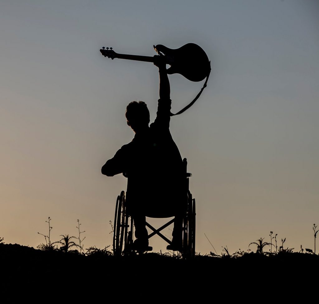 Silhouette of a person in a wheelchair on a hilltop at sunset raising a guitar in triumph.