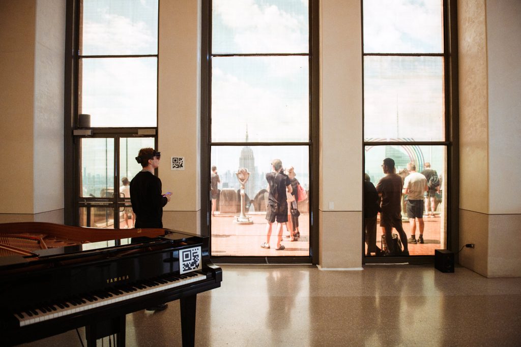 View from inside of the observation deck of the Top of the Rockefeller Center. People are casually walking around and inside there is a grand piano.