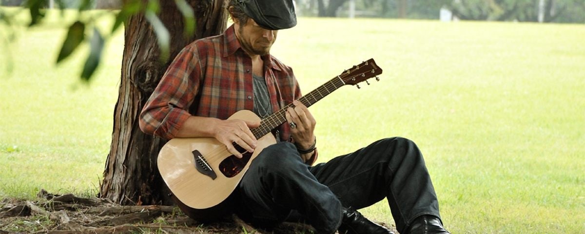 Young man sitting under a tree playing an acoustic guitar.