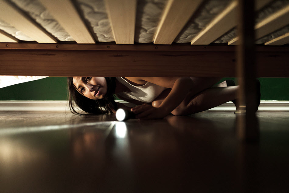 Young woman using a flashlight to look under the bed.