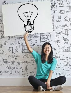 woman sitting in front of wall full of drawn doodles and holding up card with a drawing of a lightbulb