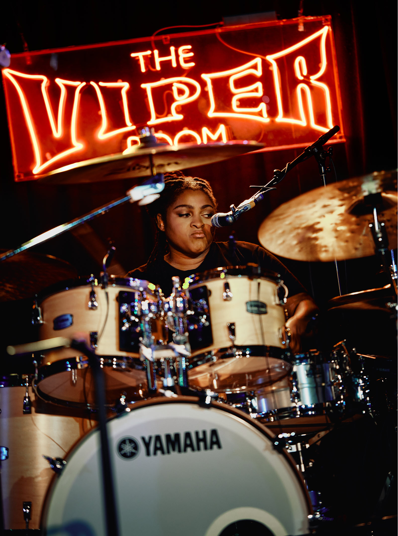 Woman playing drums with lit up sign behind her that says "The Viper Room".
