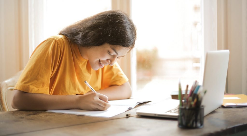 teen girl writing in notebook with open laptop in front of her