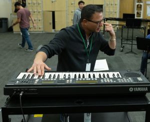 music educator at keyboard and pointing to his ear