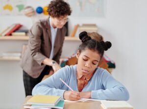 teacher working with a student in a classroom while another students works on a worksheet