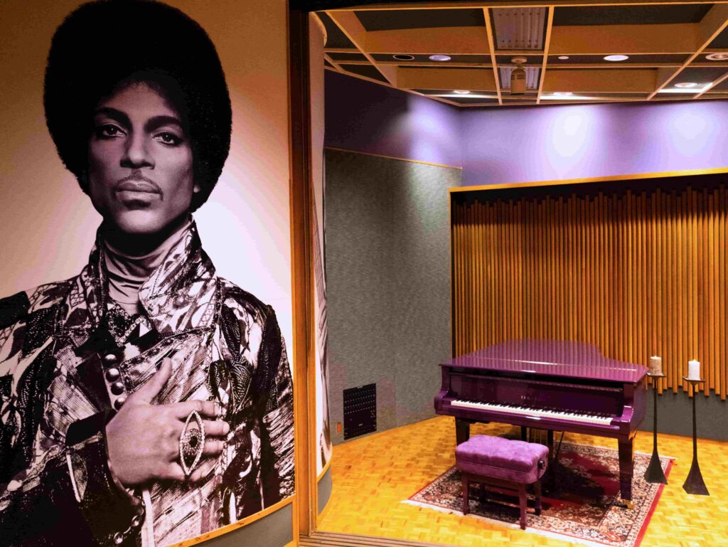 A purple piano in a recording studio with a poster of Prince on the wall.