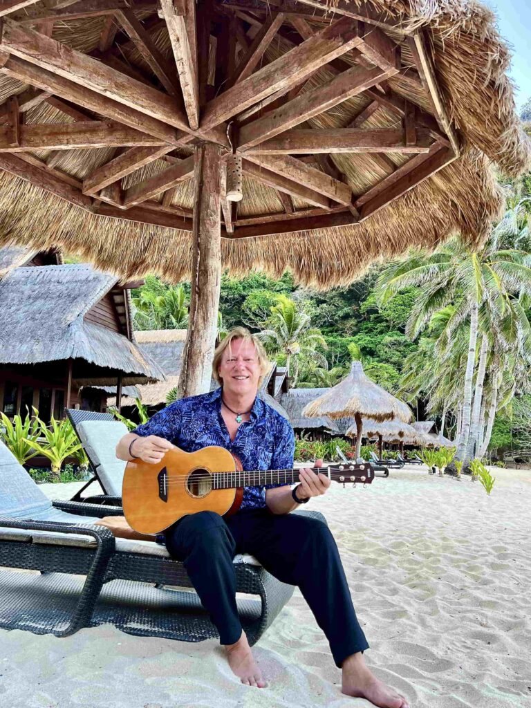 A smiling man on a beach playing an acoustic guitar.