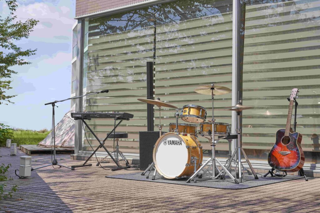 A boom microphone, electronic keyboard, P.A. system, acoustic drum kit and acoustic guitar set up outside an office building.