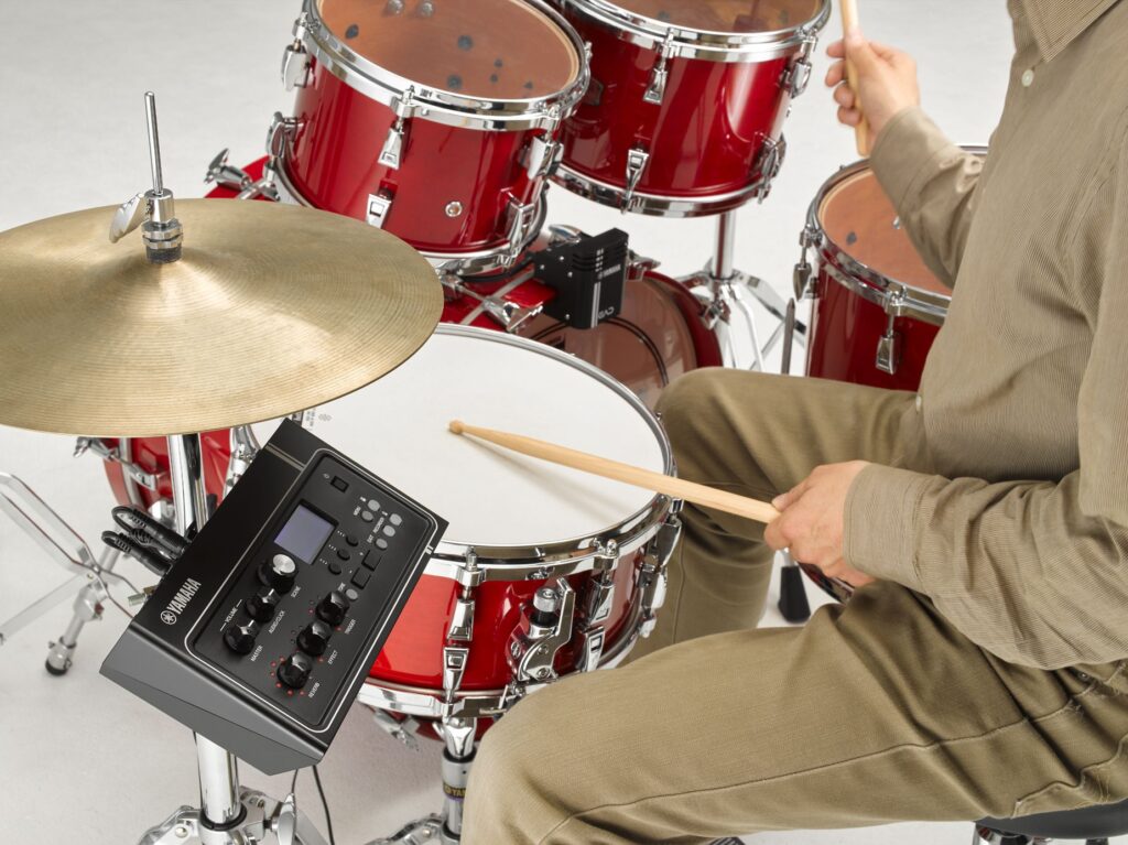 Man playing a red drum kit with a sensor attached to the bass drum and a module mounted below the hi-hat cymbals.