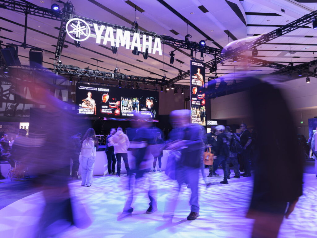 People walking by the main stage in the Yamaha exhibit at 2025 NAMM, some of them blurred.