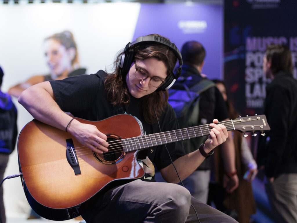 A smiling young man wearing headphones playing an acoustic guitar at 2025 NAMM.