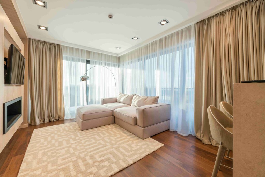 A room with a throw rug, a sofa, and curtains on two large windows.