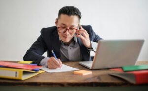 busy man holding phone and writing on paper with an open laptop in front of him