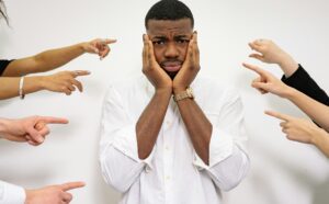 man holding face in hands while several hands are pointing at him 