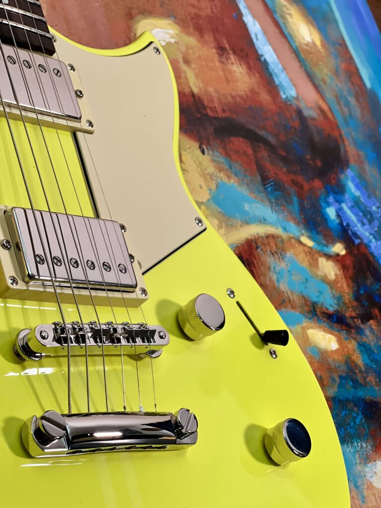 A closeup of a yellow electric guitar bridge showing two pickups, two knobs and a pickup selector switch.