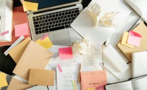 messy desk with laptop, notebooks, crumbled paper strewn everywhere