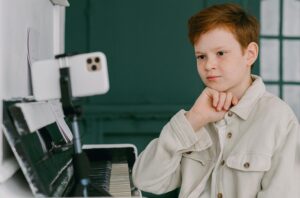 young male student sitting at piano with mobile phone pointing at him