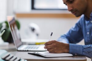 man sitting in front of laptop and writing in notebook 