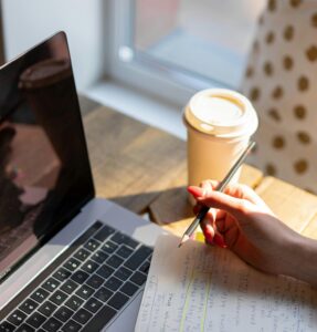 someone taking notes with open laptop and coffee on desk