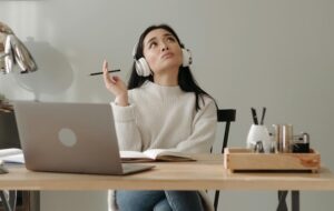 thinking woman wearing headphones and sitting at desk with open laptop
